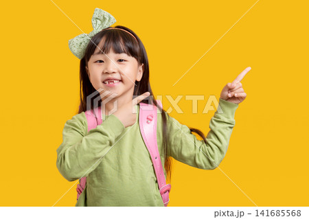 Cute Asian schoolgirl pointing sideways with finger, smiling child with backpack on yellow background, advertising and direction concept 141685568