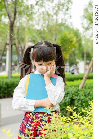 A thoughtful young student holding a blue folder and looking down while standing next to green bushes. 141685678