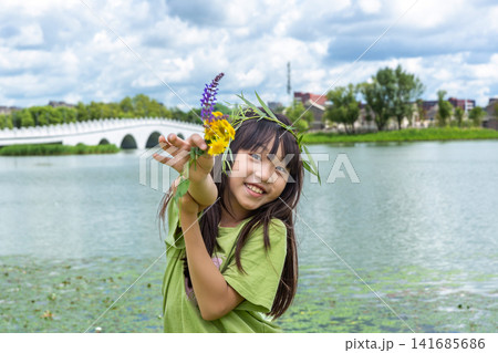 A young girl wearing a handmade willow garland and holding wildflowers smiles near a scenic riverbank. A young girl wearing a handmade willow garland and holding wildflowers smiles near a scenic riverbank. 141685686