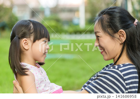 A happy mother and her little daughter smiling and looking at each other affectionately in a green park during summertime. 141686958
