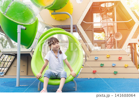 A young girl in a white t-shirt and jeans sits inside a green tunnel at a modern outdoor playground with warm sunlight. A young girl in a white t-shirt and jeans sits inside a green tunnel at a modern outdoor playground with warm sunlight. 141688064