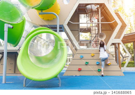 An active little girl climbs a hexagonal bouldering wall at a colorful outdoor playground under bright sunlight. 141688065