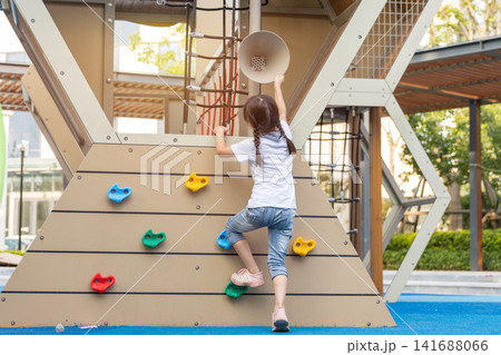 Back view of a brave young girl reaching for the top of a modern climbing frame at a neighborhood park. 141688066