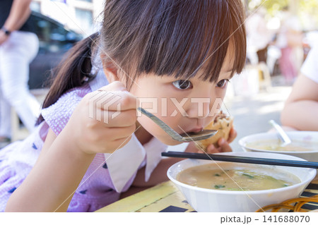 A focused little girl in a purple dress uses a spoon to eat a delicious traditional breakfast at an outdoor food stall. A focused little girl in a purple dress uses a spoon to eat a delicious traditional breakfast at an outdoor food stall. 141688070