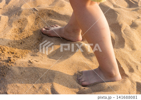Close-up detail of a child's bare feet walking on soft golden sand at the beach during sunset. Close-up detail of a child's bare feet walking on soft golden sand at the beach during sunset. 141688081