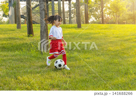 Active little girl in red and white uniform running after a soccer ball on green grass hill. Active little girl in red and white uniform running after a soccer ball on green grass hill. 141688266