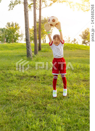 Enthusiastic young goalkeeper wearing gloves and catching a soccer ball in mid-air at the park. 141688268