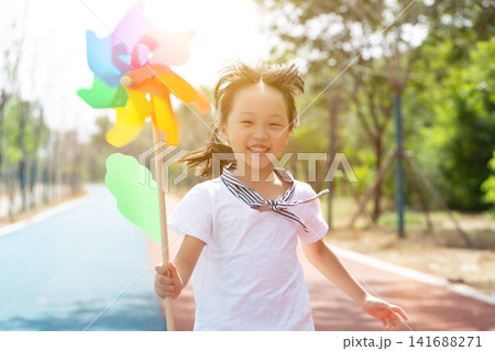 Cheerful little girl running forward while holding a colorful pinwheel toy on a park path. 141688271
