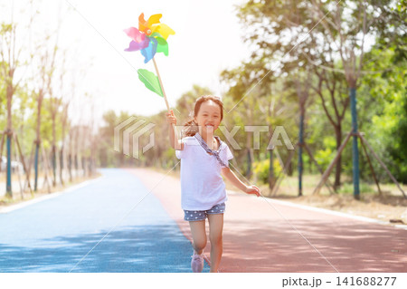 Cheerful little girl running forward while holding a colorful pinwheel toy on a park path. Cheerful little girl running forward while holding a colorful pinwheel toy on a park path. 141688277