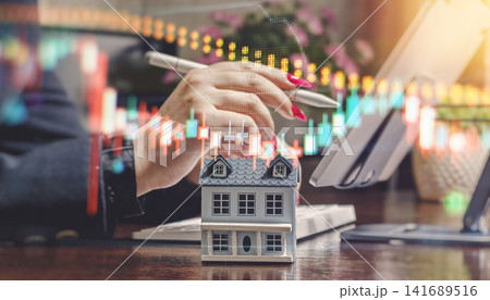 Real estate market volatility and investment concept. Miniature house with digital financial candlestick charts and businesswoman working in background. Real estate market volatility and investment concept. Miniature house with digital financial candlestick charts and businesswoman working in background. 141689516