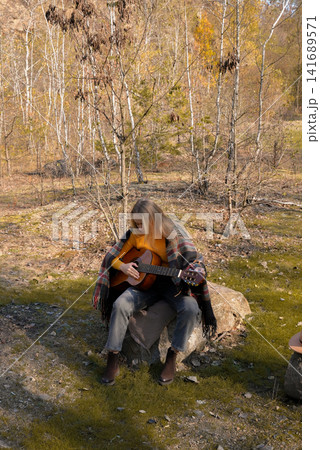 Beautiful blonde girl playing acoustic guitar and singing relaxing enjoying outdoors on sunny autumn day. Hippie woman in boho style learning and practicing playing musical instrument. Fall leaves Beautiful blonde girl playing acoustic guitar and singing relaxing enjoying outdoors on sunny autumn day. Hippie woman in boho style learning and practicing playing musical instrument. Fall leaves 141689571