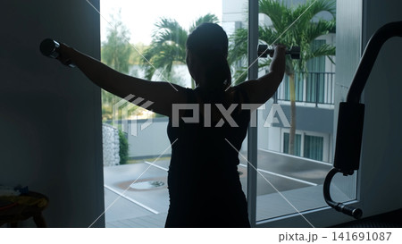 Dark silhouette of young woman performing lateral dumbbell raises with both arms extended sideways in a gym with bright window and tropical palms behind. Fitness concept Dark silhouette of young woman performing lateral dumbbell raises with both arms extended sideways in a gym with bright window and tropical palms behind. Fitness concept 141691087