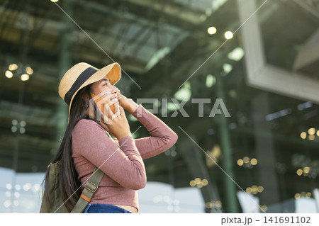 Happy Asian woman tourist smile with phone calling, Backpack female traveler enjoy journey trip tour standing against airport building facade with smartphone 141691102