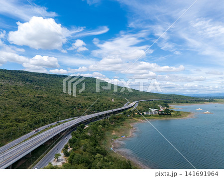 Drone aerial view of a highway running beside a reservoir with lush green mountains, Transportation infrastructure and renewable energy concept Drone aerial view of a highway running beside a reservoir with lush green mountains, Transportation infrastructure and renewable energy concept 141691964