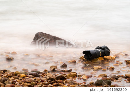 Professional camera resting pebbles beside calm water captured long exposure creating smooth motion ideal for travel photography nature landscape outdoor themes Professional camera resting pebbles beside calm water captured long exposure creating smooth motion ideal for travel photography nature landscape outdoor themes 141692290