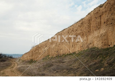 Long eroded clay cliff with bird nesting holes along a dirt path in Visnjicka Banja, Belgrade. Natural geological wall under soft cloudy sky Long eroded clay cliff with bird nesting holes along a dirt path in Visnjicka Banja, Belgrade. Natural geological wall under soft cloudy sky 141692409