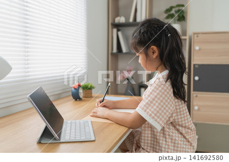 Young girl sitting at desk writing with pen using digital tablet with keyboard in bright modern room 141692580