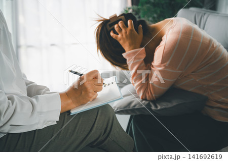 Distressed female client covers head during counseling while therapist writes notes, showing stress, anxiety, emotional support, mental health care, empathy, and private therapy indoors. 141692619