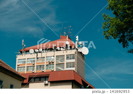 A city building rooftop is covered with satellite dishes and communication antennas under a clear blue sky. Urban technology, telecom infrastructure, broadcasting, and digital connectivity A city building rooftop is covered with satellite dishes and communication antennas under a clear blue sky. Urban technology, telecom infrastructure, broadcasting, and digital connectivity 141692953