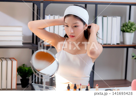 Young asian woman wearing headband and tying hair while preparing skincare looking mirror in room. 141693428