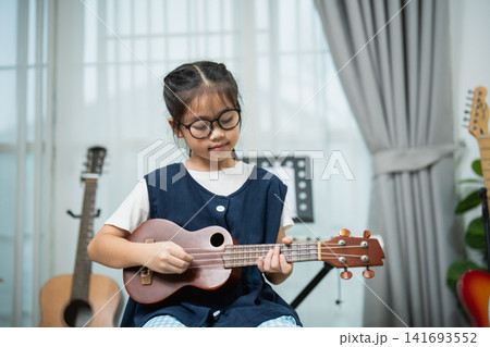 Asian Little Girl with Glasses Playing Ukulele at Home with Acoustic Guitar and Musical Instruments Background 141693552
