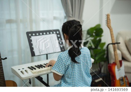 Back View of Little Asian Girl Playing Electric Keyboard at Home with Sheet Music and Guitars in Cozy Living Room Back View of Little Asian Girl Playing Electric Keyboard at Home with Sheet Music and Guitars in Cozy Living Room 141693569