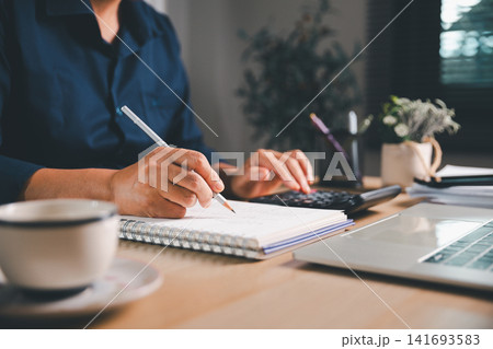 Business professional writing financial notes and using a calculator beside a laptop and coffee cup, representing accounting, bookkeeping, budget planning, and expense management in a home office. Business professional writing financial notes and using a calculator beside a laptop and coffee cup, representing accounting, bookkeeping, budget planning, and expense management in a home office. 141693583