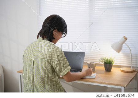 Rear view of Asian woman taking notes on notepad while working on laptop at wooden desk near window in bright home office workspace 141693674