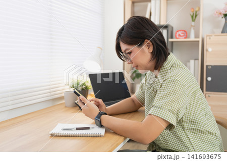 Asian woman using smartphone while sitting at wooden desk with laptop in bright minimalist home office workspace Asian woman using smartphone while sitting at wooden desk with laptop in bright minimalist home office workspace 141693675