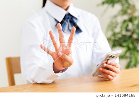 リビングのテーブルでスマホを見て指を4本立てる制服姿の中学生 リビングのテーブルでスマホを見て指を4本立てる制服姿の中学生 141694169