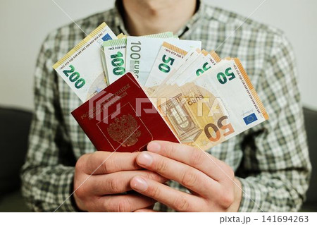 Hands hold a Russian passport with mixed euro banknotes in front of the body. Concept of relocation from Russia, migration budget and financial planning for moving abroad 141694263