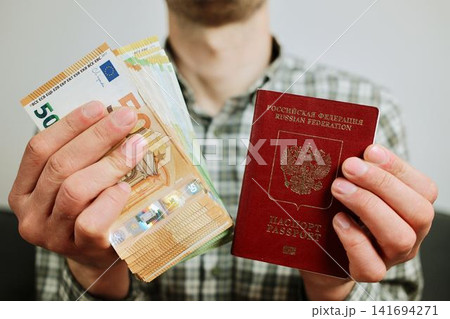 Hands hold a Russian passport with mixed euro banknotes in front of the body. Concept of relocation from Russia, migration budget and financial planning for moving abroad Hands hold a Russian passport with mixed euro banknotes in front of the body. Concept of relocation from Russia, migration budget and financial planning for moving abroad 141694271