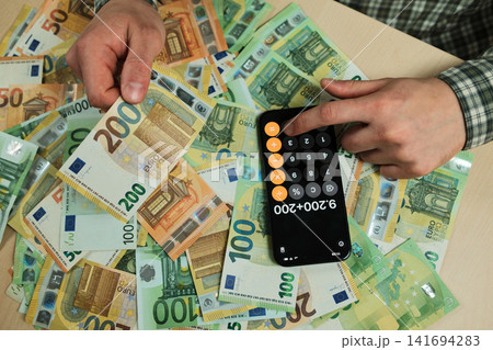 Hand holds a two hundred euro banknote above scattered cash beside a smartphone calculator. Concept of wealth, business finance and payment planning for banking or economy themes 141694283