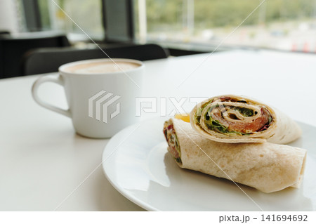 Salmon tortilla wrap with cappuccino on a bright cafe table near the window. Concept of urban breakfast, business lunch, coffee culture and restaurant branding Salmon tortilla wrap with cappuccino on a bright cafe table near the window. Concept of urban breakfast, business lunch, coffee culture and restaurant branding 141694692
