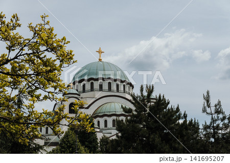 Saint Sava Temple rises above green trees under cloudy sky in Belgrade Serbia. Concept of spirituality, tourism, heritage and urban nature Saint Sava Temple rises above green trees under cloudy sky in Belgrade Serbia. Concept of spirituality, tourism, heritage and urban nature 141695207