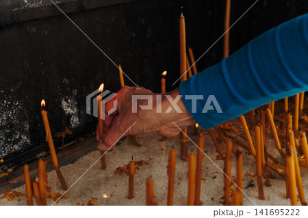 Person places a lit candle into white sand inside a religious shrine. Concept of prayer, faith, spirituality and hope Person places a lit candle into white sand inside a religious shrine. Concept of prayer, faith, spirituality and hope 141695222