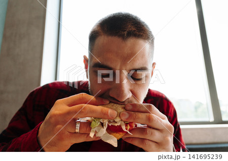 Young man eating a fresh burger near window light in modern cafe interior. Concept of fast food lifestyle comfort hunger lunch and casual urban dining Young man eating a fresh burger near window light in modern cafe interior. Concept of fast food lifestyle comfort hunger lunch and casual urban dining 141695239