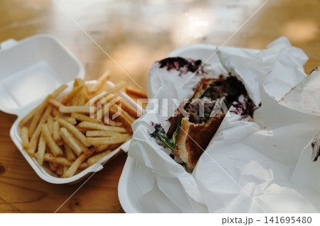 Burger with french fries served in takeaway packaging on wooden table. Concept of street food, fast food and casual dining Burger with french fries served in takeaway packaging on wooden table. Concept of street food, fast food and casual dining 141695480