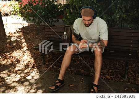 Young man sitting on a park bench with headphones, checking smartwatch. Concept of relaxation, health tracking, and lifestyle Young man sitting on a park bench with headphones, checking smartwatch. Concept of relaxation, health tracking, and lifestyle 141695939