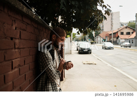 Young man leaning against a brick wall on a city street, checking the time on his watch. Concept of punctuality, waiting, and urban lifestyle Young man leaning against a brick wall on a city street, checking the time on his watch. Concept of punctuality, waiting, and urban lifestyle 141695986