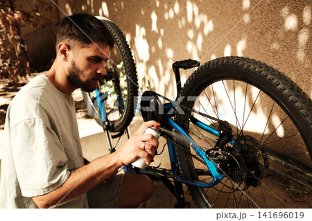 Focused man maintaining his blue mountain bike by spraying lubricant on the chain outdoors. Concept of care, maintenance, and active living Focused man maintaining his blue mountain bike by spraying lubricant on the chain outdoors. Concept of care, maintenance, and active living 141696019