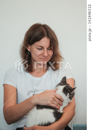 Woman gently holding a gray and white domestic cat indoors. Calm interaction reflects affection, trust, and responsible pet ownership 141696318