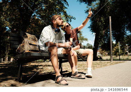 Young man and woman sitting on a park bench eating ice cream while the woman points at something. Concept of friendship, joy, and summer freedom Young man and woman sitting on a park bench eating ice cream while the woman points at something. Concept of friendship, joy, and summer freedom 141696544