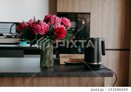 Stylish modern kitchen interior with bouquet of pink peonies in glass vase and electric kettle on dark countertop. Stylish modern kitchen interior with bouquet of pink peonies in glass vase and electric kettle on dark countertop. 141696849