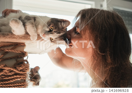 Woman sharing an affectionate moment by kissing her domestic cat resting on a cat tree. Warm indoor lifestyle scene highlighting love, companionship, trust, and pet care 141696928