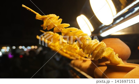 Crispy spiral potato skewer served in clay pot with warm market bokeh, street food concept Crispy spiral potato skewer served in clay pot with warm market bokeh, street food concept 141697151