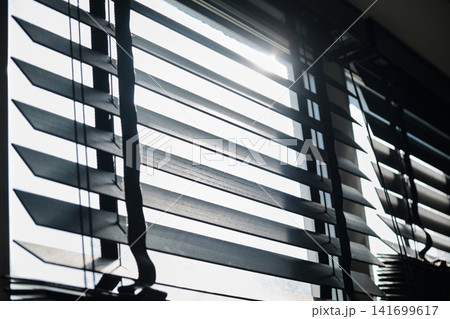 Low-angle view of horizontal wooden venetian window blinds against bright backlighting and lens flare. Sunlight streams directly through the open slats, creating a warm and bright ambiance Low-angle view of horizontal wooden venetian window blinds against bright backlighting and lens flare. Sunlight streams directly through the open slats, creating a warm and bright ambiance 141699617