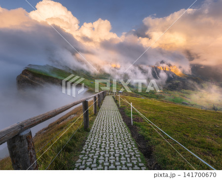 Path with wooden fence and mountain cliffs in clouds at sunset Path with wooden fence and mountain cliffs in clouds at sunset 141700670
