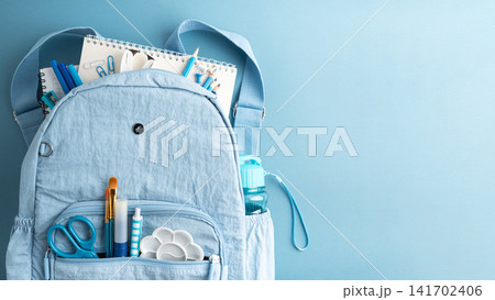 Light blue backpack filled with school and art supplies on a light blue background 141702406
