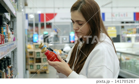 Woman making purchasing decision in supermarket, holding two bottles of tomato sauce and juice, reading labels, surrounded by various products on shelves Woman making purchasing decision in supermarket, holding two bottles of tomato sauce and juice, reading labels, surrounded by various products on shelves 141705267